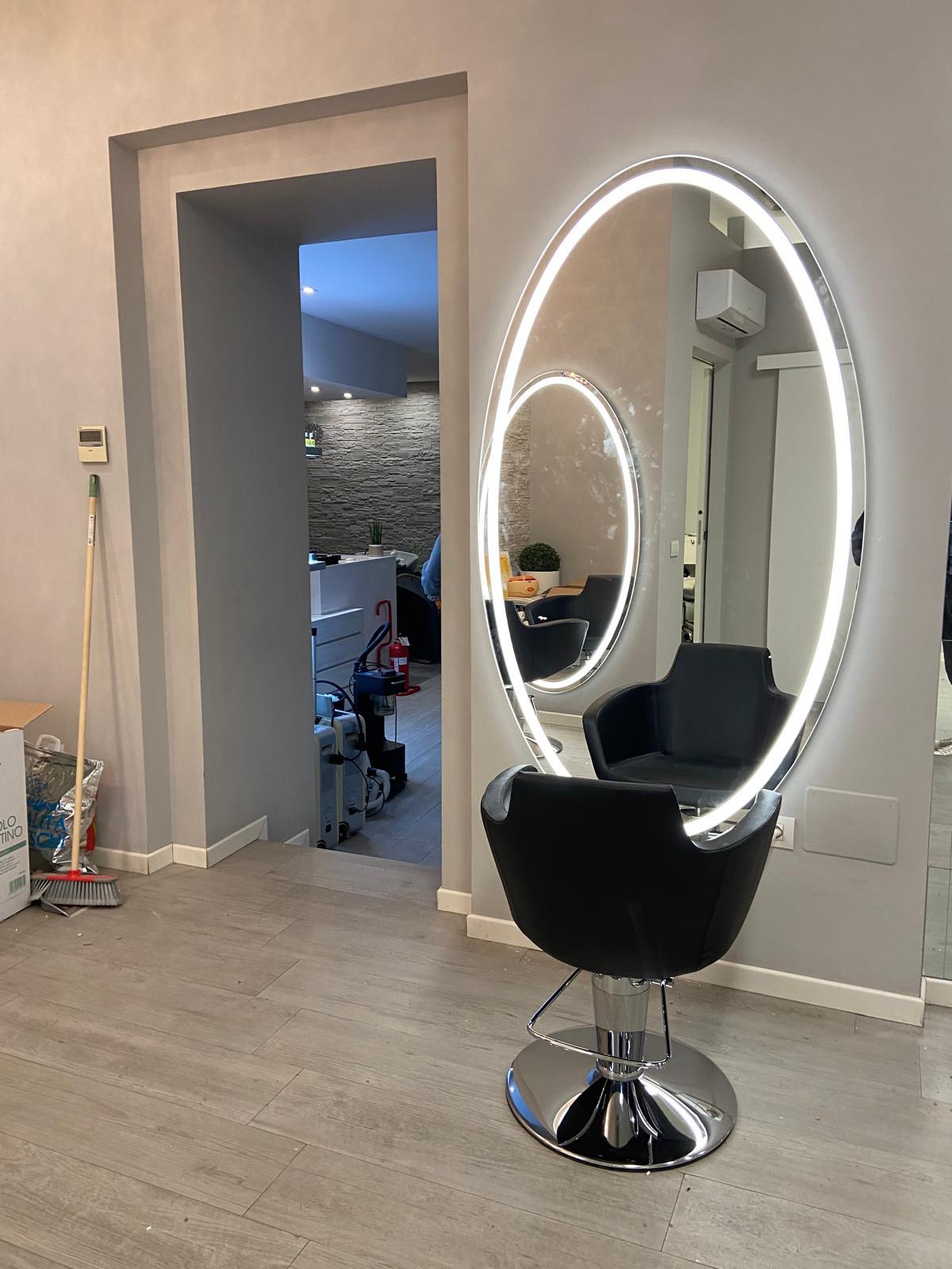 Styling area with a black salon chair positioned before a large illuminated oval mirror, with another room visible through the doorway.
