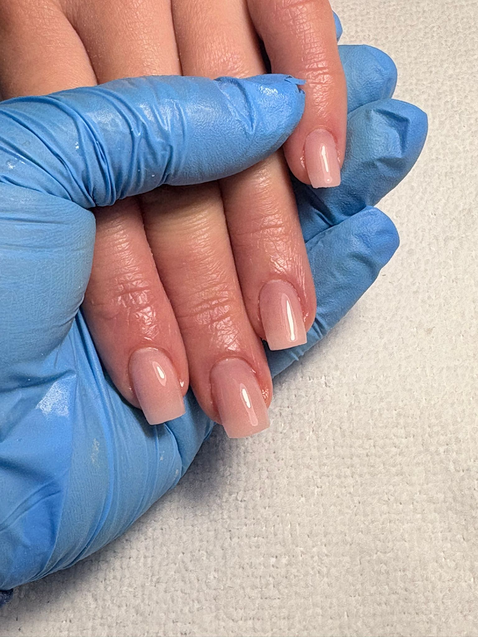 Close-up of manicured fingers being held by blue-gloved hands, with glossy pale-pink nails against a light textured surface.