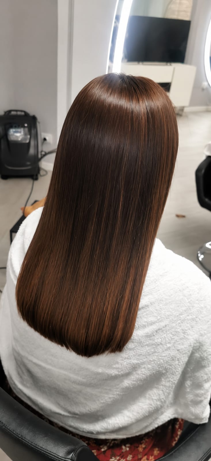 Rear view of a seated person with long, straight brown hair, framed in a salon chair under bright indoor lighting.