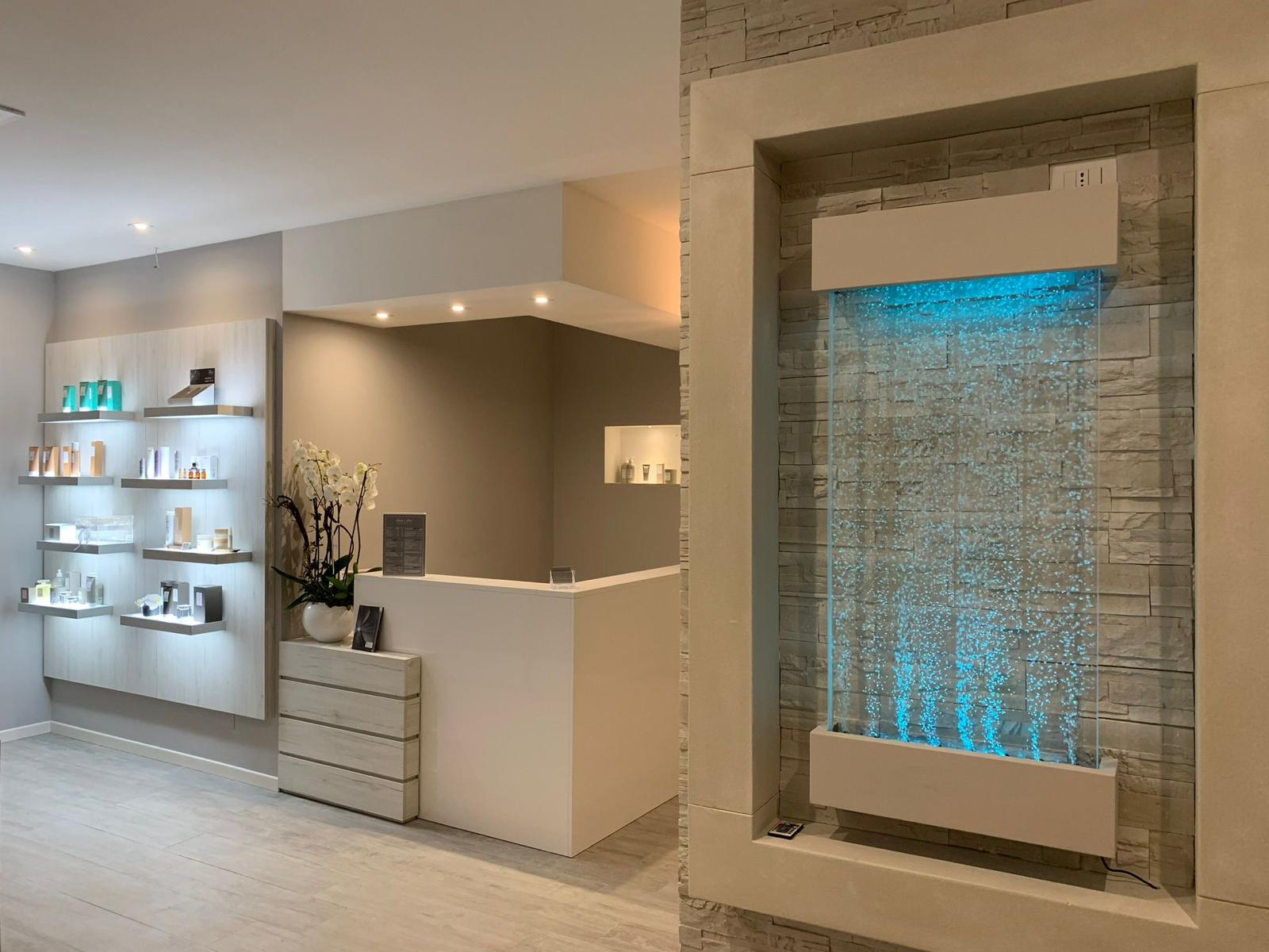 Salon reception area with illuminated product shelving, a white counter and a blue-lit wall fountain, framed in a bright modern interior.