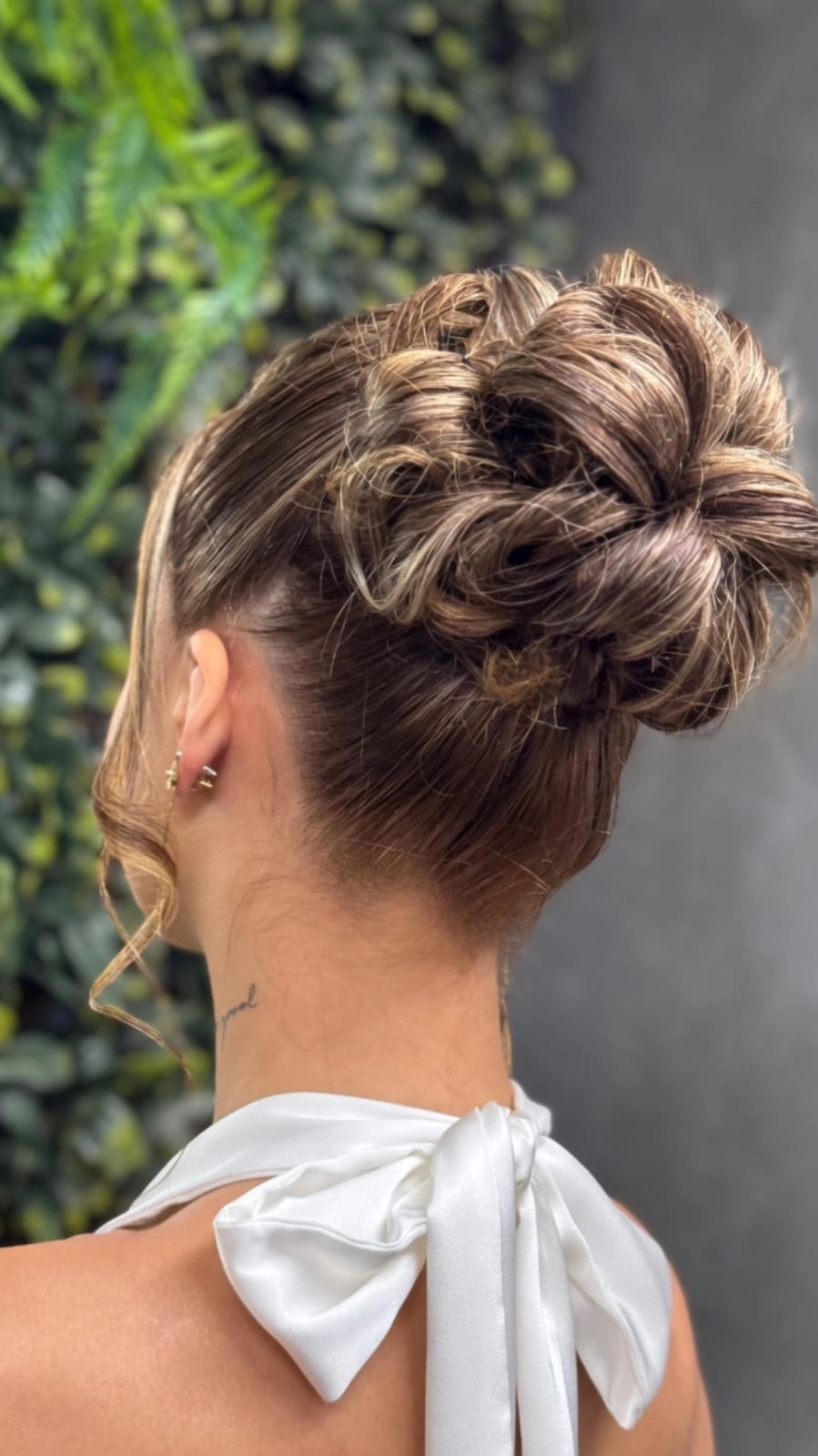 Close-up rear view of a dark-haired high bun with loose tendrils, against a softly lit leafy background.