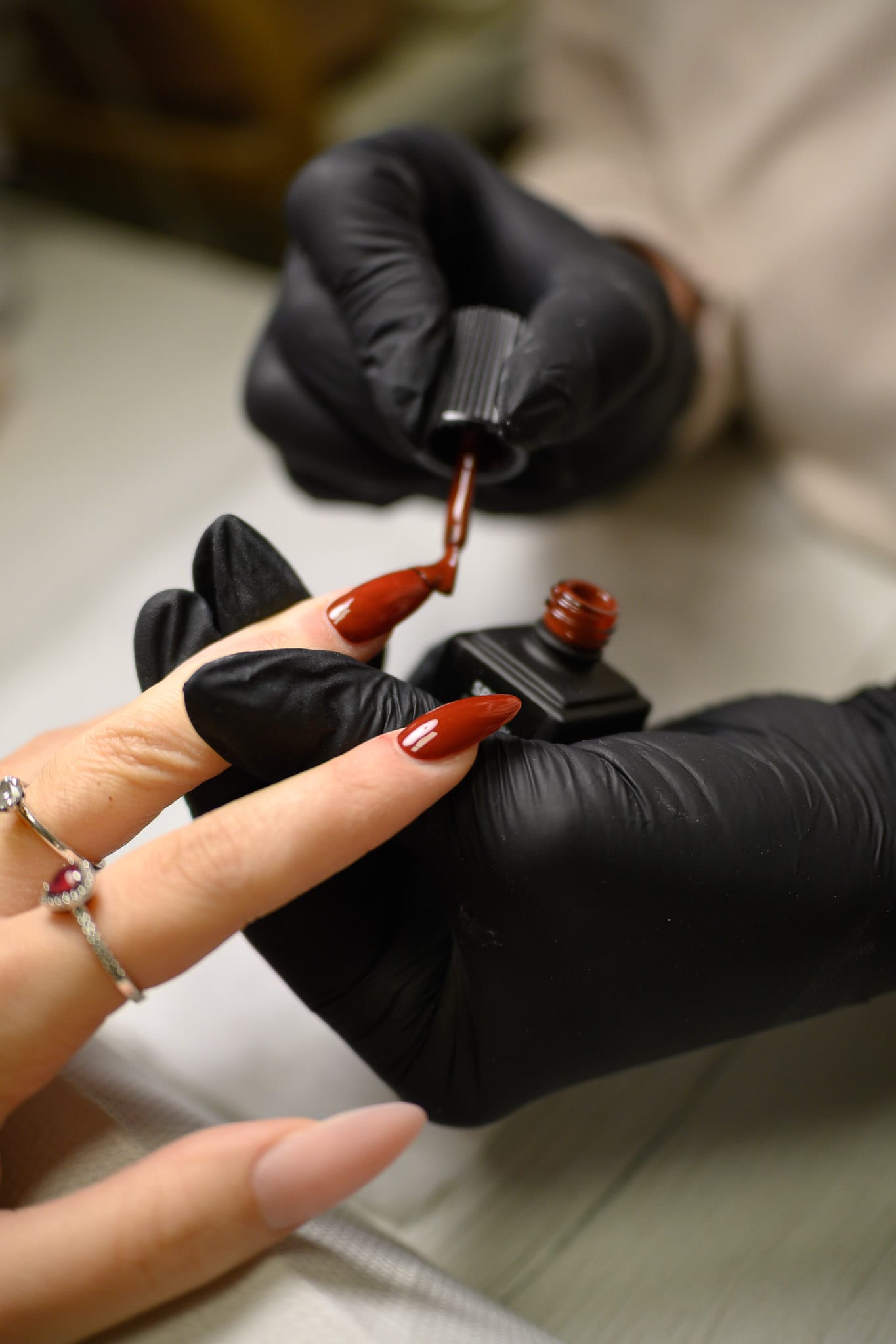 Close-up of black-gloved hands applying red polish to a fingernail with a brush beside an open polish bottle.