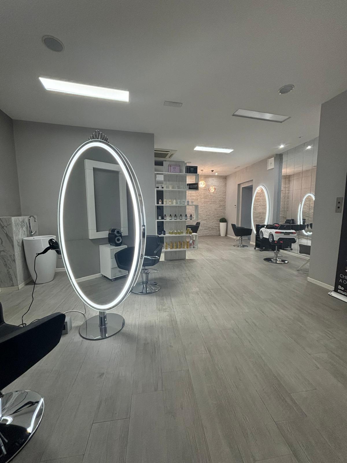 Wide salon interior with illuminated oval mirrors, styling chairs, product shelving and bright rectangular ceiling lights.