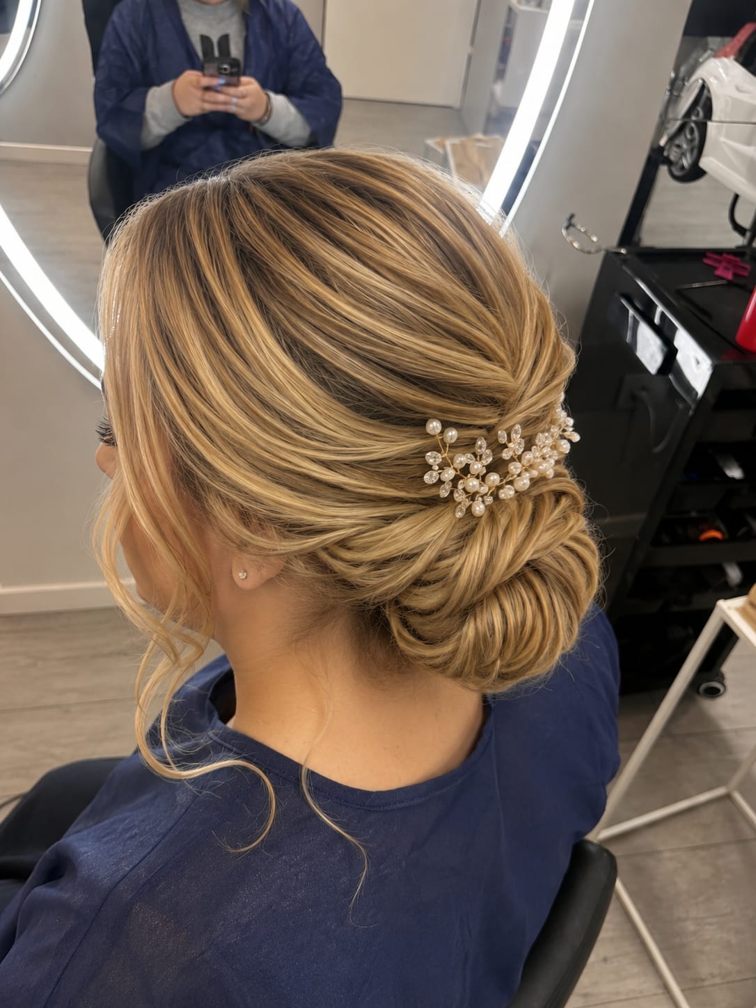 Close-up rear view of a blonde low updo with a jeweled hair accessory, seated in a salon chair under bright ring lighting.